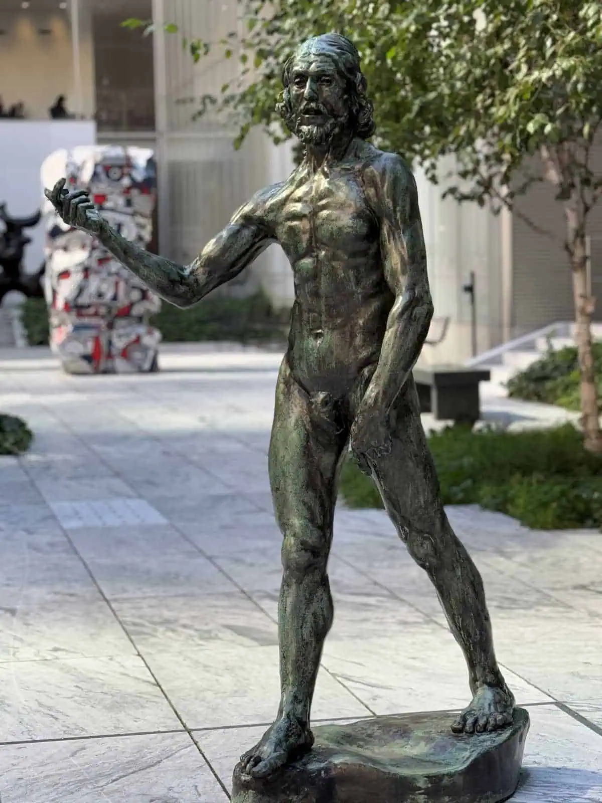 New York City Before A Cruise: Sailing From the Red Hook Cruise Terminal 3 Bronze statue of an ancient Greek man holding a scroll, located outdoors in an urban setting with trees and modern buildings in the background.