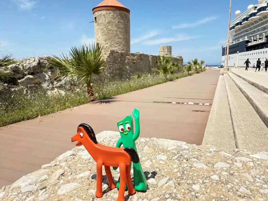 1 Day in Port: Exploring Rhodes Old Town on Foot – A Perfect Self-Guided Walk 16 Colorful toy figures of Gumby & Pokey on a stone wall at Rhodes port, with a historic tower and cruise ship in the background.