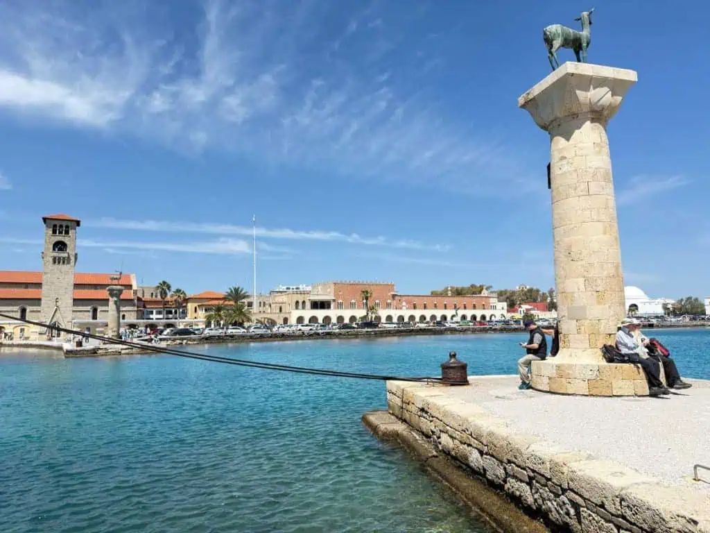 1 Day in Port: Exploring Rhodes Old Town on Foot – A Perfect Self-Guided Walk 8 Scenic view of Rhodes Old Town harbor with historic buildings, a deer statue on a column, and people relaxing by the water on a sunny day.