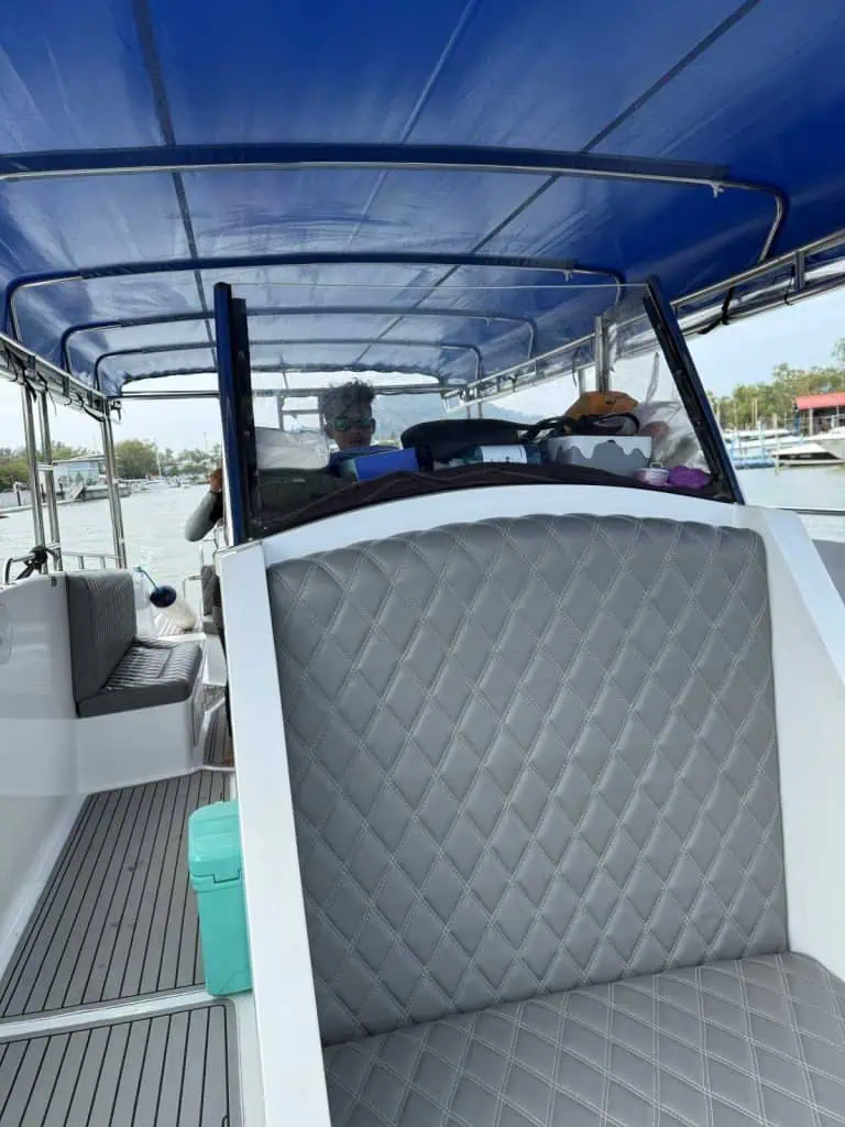 1 Day in Port: Phuket – A Private Phang Nga Bay Tour By Speedboat 3 Phuket – A Private Phang Nga Bay Tour By Speedboat Interior view of a speedboat in Phuket with cushioned seats and a blue canopy, on a calm waterway with boats and trees in the background.